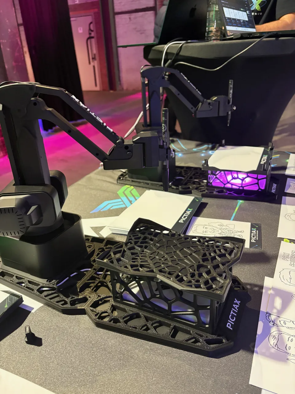 PictiaX® robot arms on the booth table, with a base lit in purple and caricatures lying on the table